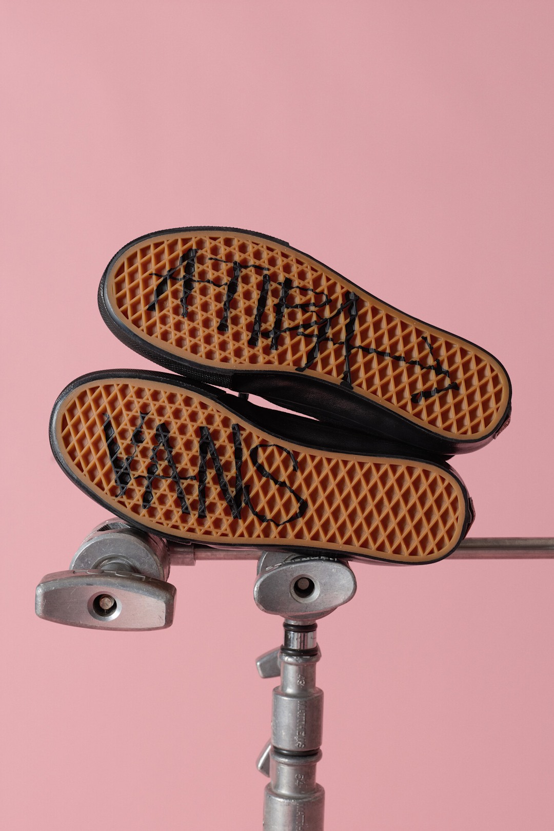 A pair of Vans Authentic shoes with waffle-pattern outsoles visible are balanced on a skateboard truck against a soft pink background. 