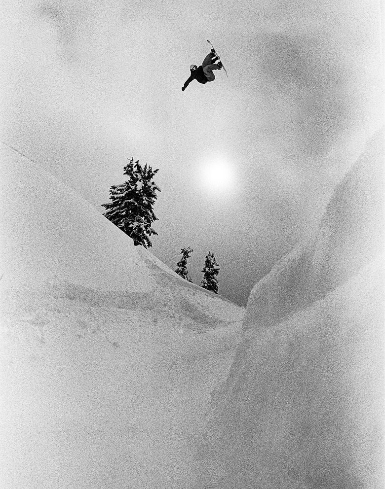 A grainy black and white photo of a snowboarder doing a trick in the air over snowy hills.
