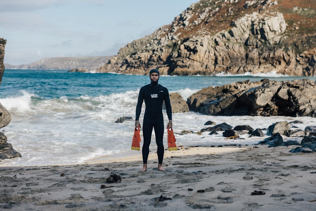 Tom Lowe in a black Vans hooded wetsuit holding orange swim fins on a rocky beach