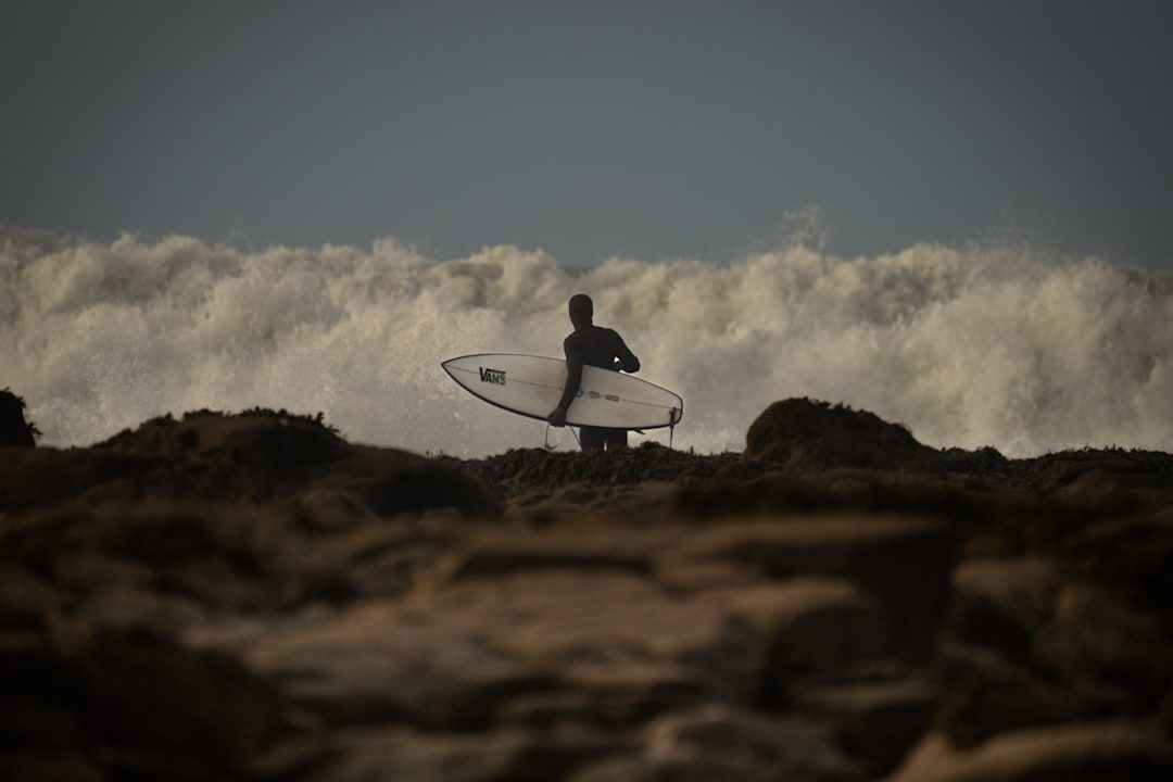 Vans surfer Tom Lowe silhouetted on the rocks with his board under his arm, facing a wall of whitewater