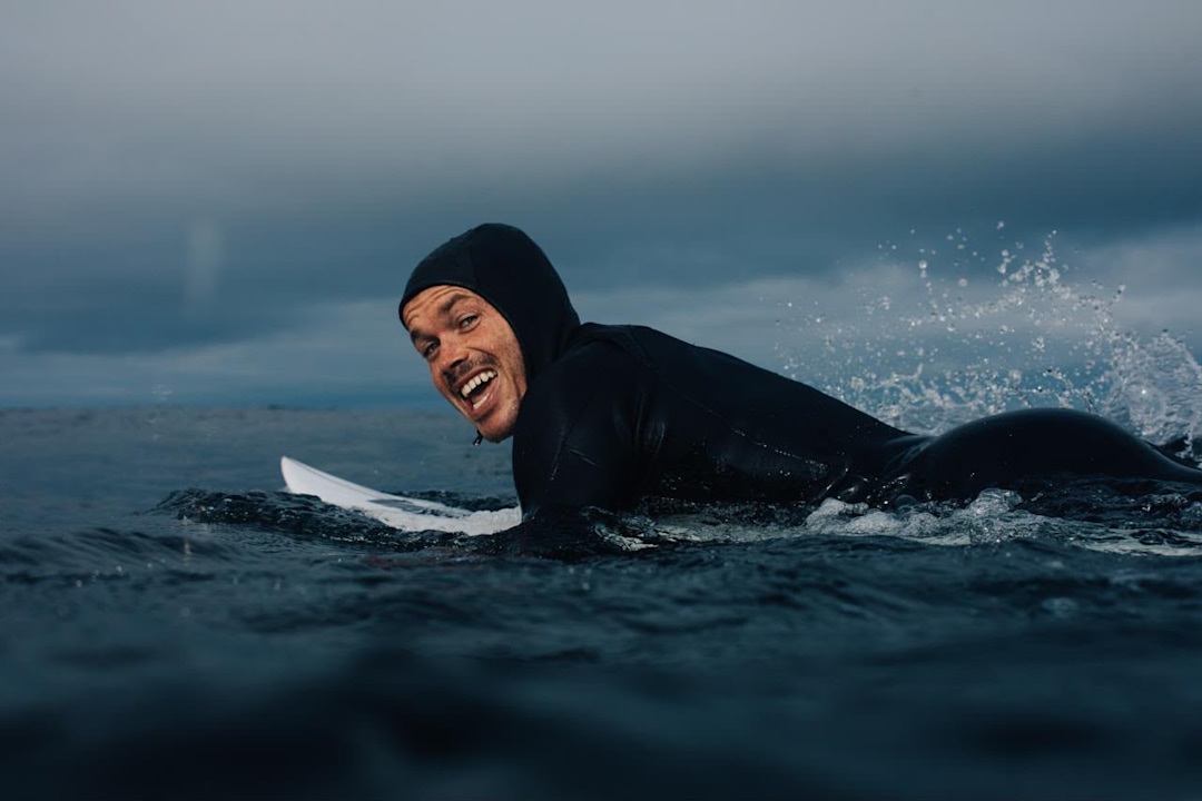Tom Lowe laying down on his surfboard in the ocean while smiling at the camera.