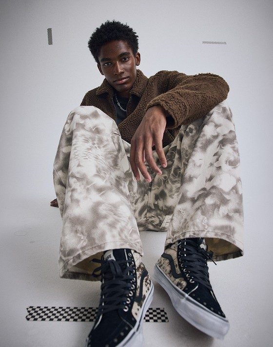 A young man sits on the floor of a photo studio wearing turtledove white and walnut brown Check-5 Baggy Printed Denim Pants.