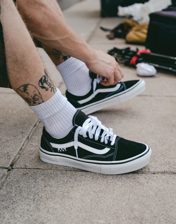 A man ties his Skate Old Skool shoes with white socks and a leg tatto.