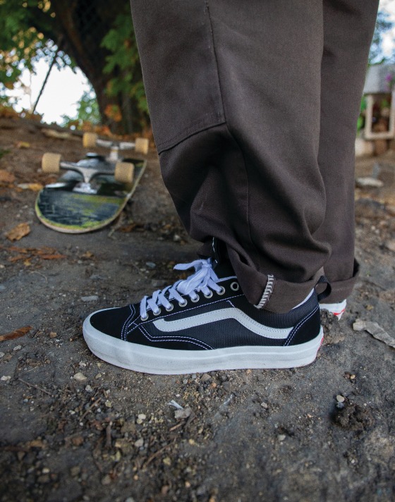 A close-up of black and gray Skate Old Skool 36+ shoes with a white Sidestripe on dirt paired with baggy charcoal pants.