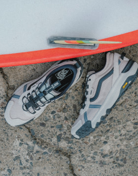 Overhead shot of white MTE Crosspath XC shoes with a blue SideStripe on the ground next to a surfboard.