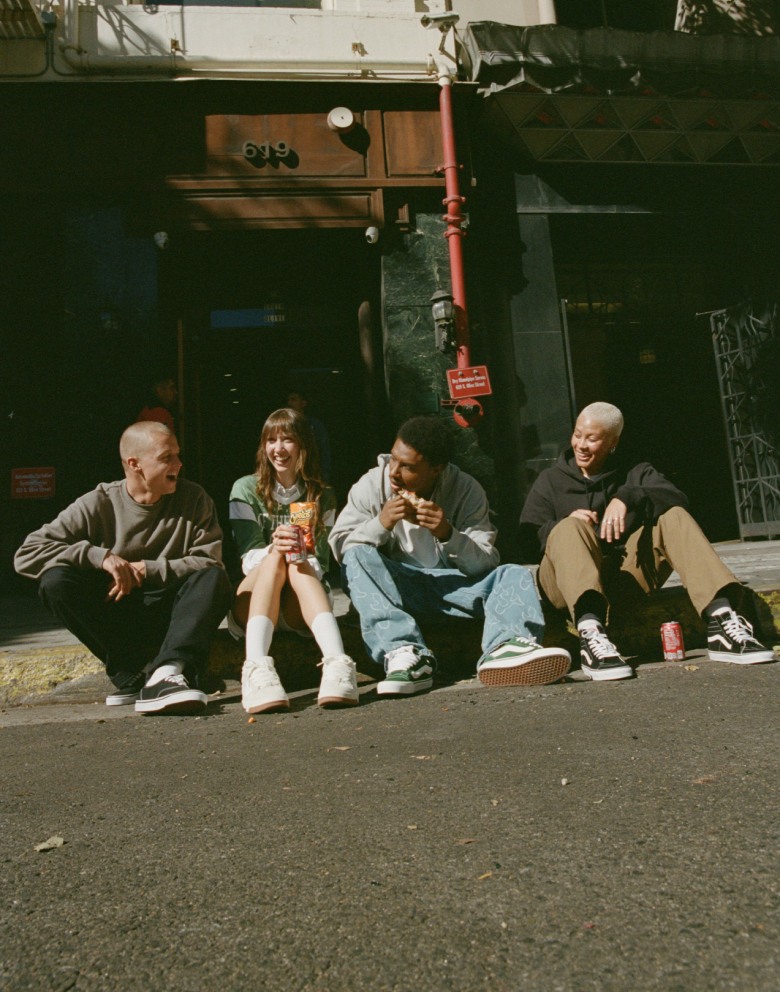 Four people sit on a sidewalk curb in Vans shoes and apparel, eating lunch.