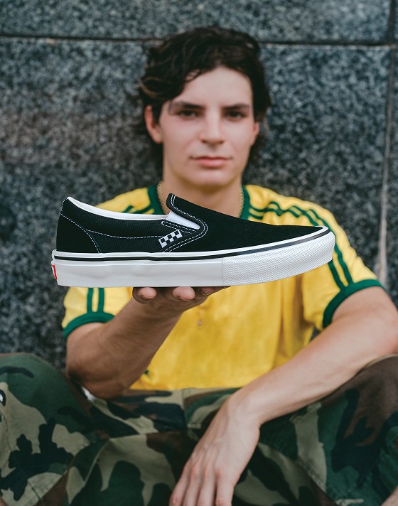Un skater professionnel accoté sur un mur porte un chandail jaune et tient des Classic Slip-On noirs à flancs blancs dans sa main.