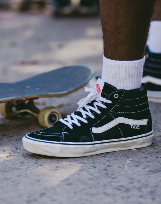A close-up of a black Sk8-Hi shoe with a white SideStripe and a skateboard behind the shoe.