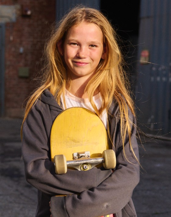 A girl with long blonde hair in a blue zip-up hoodie holding a skateboard.