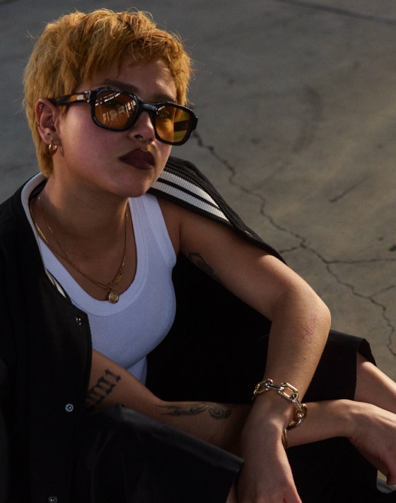 A woman with short blonde-dyed hair wearing a varsity-style jacket, a white tanktop, and sunglasses with yellow lenses.
