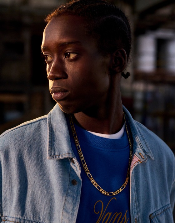 A man looking into this distance wearing a blue t-shirt with a denim button down over and a gold chain necklace.