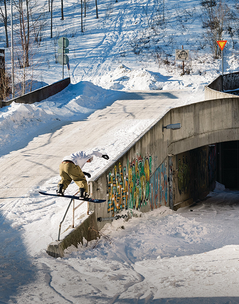 A snowboarder wearing tan snowboard pants and a white sweatshirt grinds a rail on a snow-covered road near a concrete building.