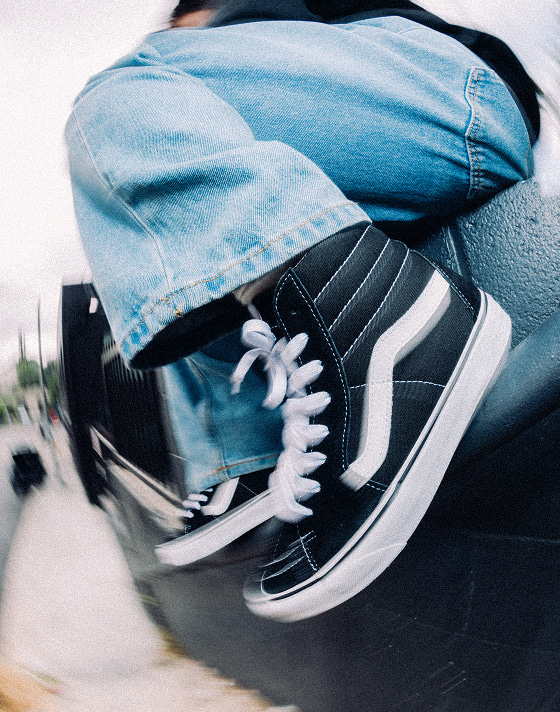 A person sitting on a handrail wearing black and white Sk8-Hi and blue jeans.