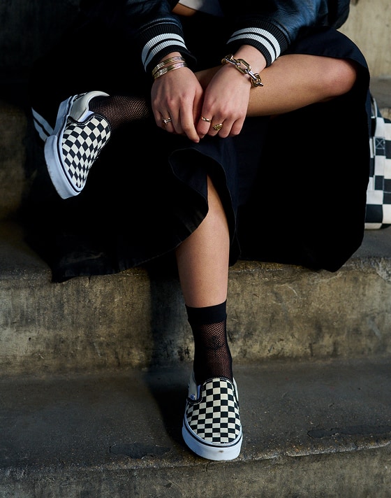A person sits on a concrete steps with black and white Checkerboard Slip-On shoes.