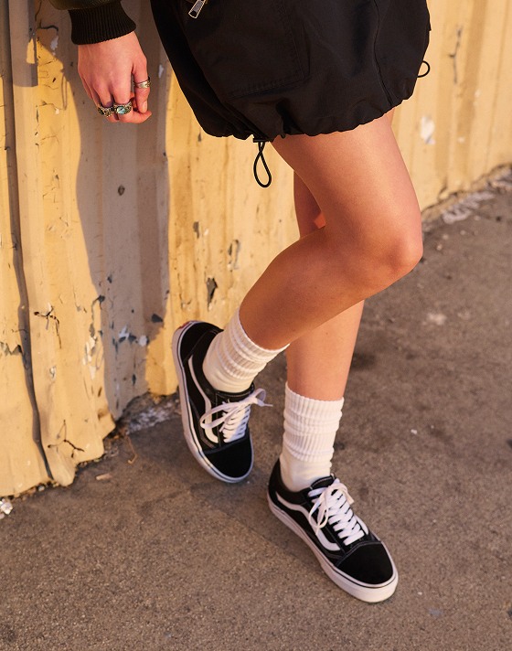 A pair of black and white Old Skool paired with white socks on a person standing on the beach.