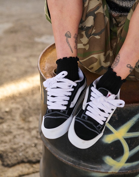 A person sitting on a metal barrel wearing black and white Knu Skool with camo shorts.