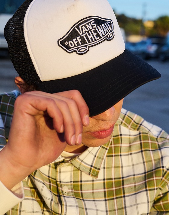 A close-up of a black and white trucker hat that says "Vans Off the Wall" with the city in the blurry background.