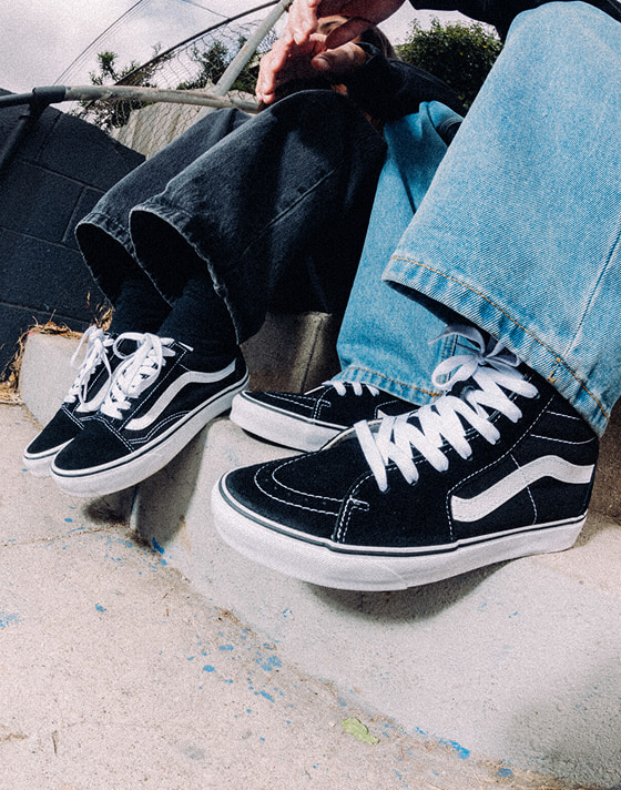 Two men sitting on concrete stairs wearing black and white Old Skool and Sk8-Hi with blue jeans.