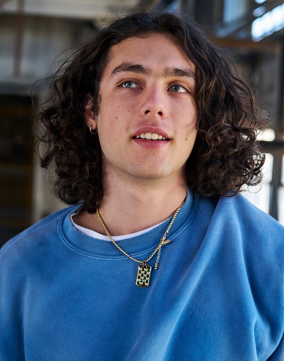 A man with long brown hair and blue eyes wearing a blue crewneck sweatshirt and a gold chain.