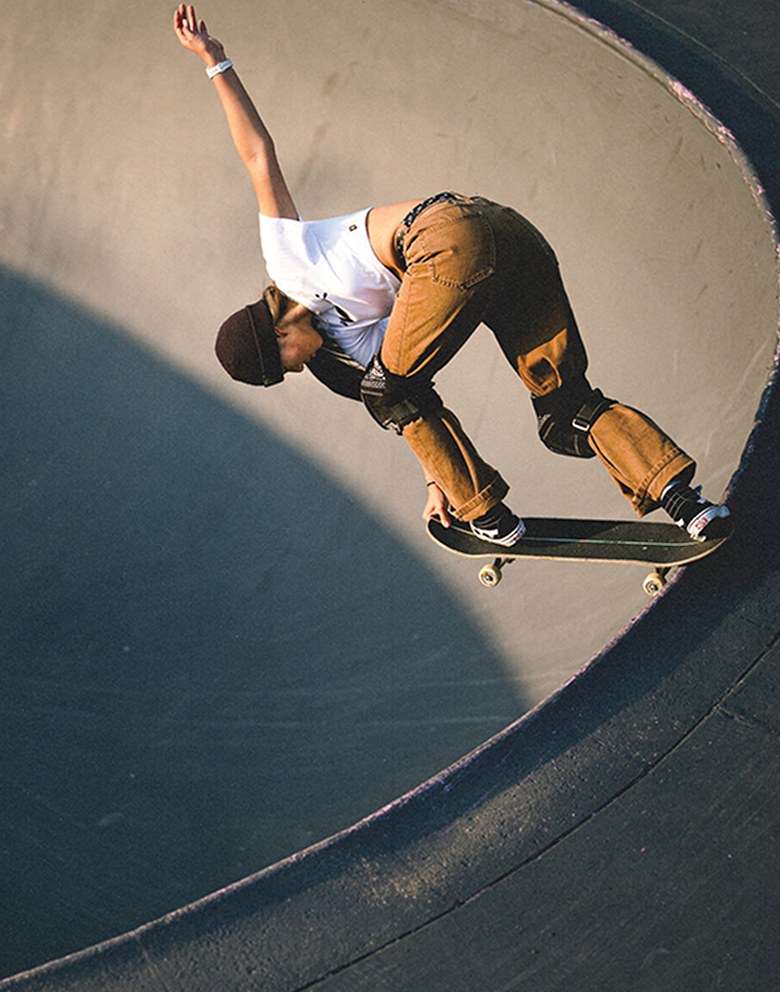 A skater grabs her board while on the lip of a bowl wearing a white t-shirt, brown pants, and black Sk8-Hi with a white SideStripe.