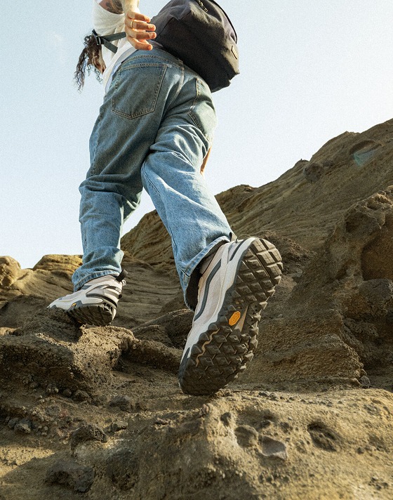 A man walking up a rocky face with white and blue MTE Crosspath shoes and blue jeans.