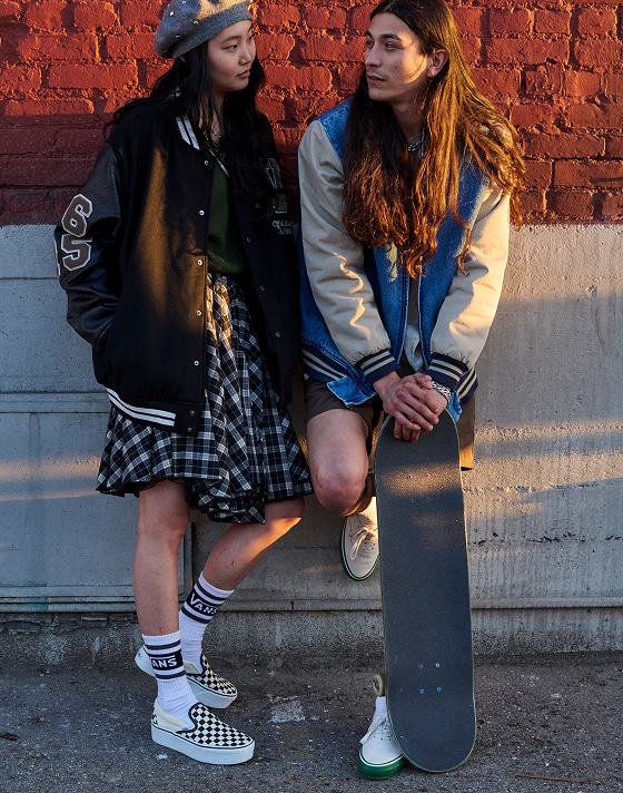 A girl in a varsity jacket and plaid skirt wearing Vans black and white checkerboard slip-on shoes standing next to a guy in a varsity jacket holding a skateboard.