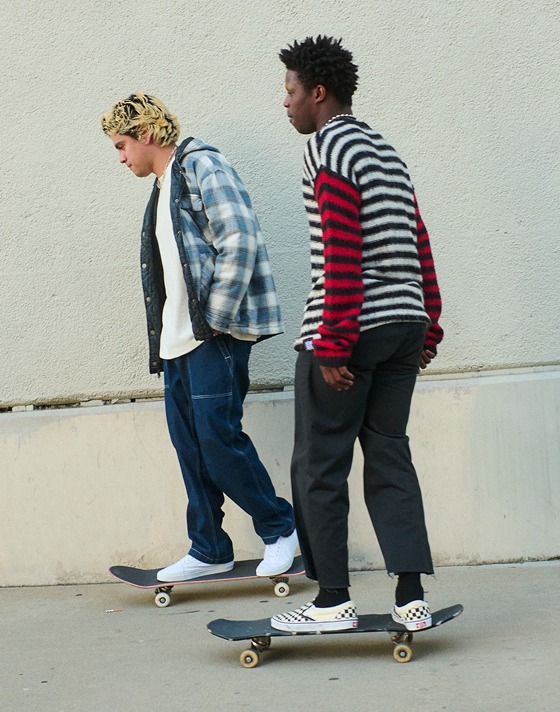 Two guys riding skateboards, one wearing Vans black and white checkerboard slip-on shoes, the other wearing white slip-on shoes.