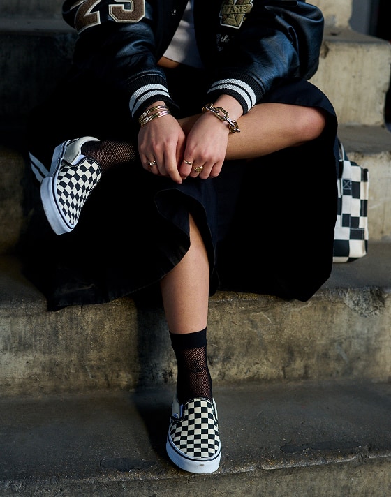 A girl sitting on concrete stairs with her leg crossed, wearing Vans black and white checkerboard slip-on shoes and Vans lacey socks.
