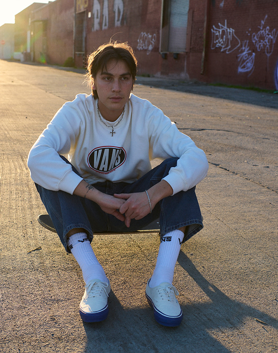 A person sitting on the ground infront of a graffiti wall, wearing a white Vans crew neck sweatshirt, white socks and white authentic shoes with blue gumsole.
