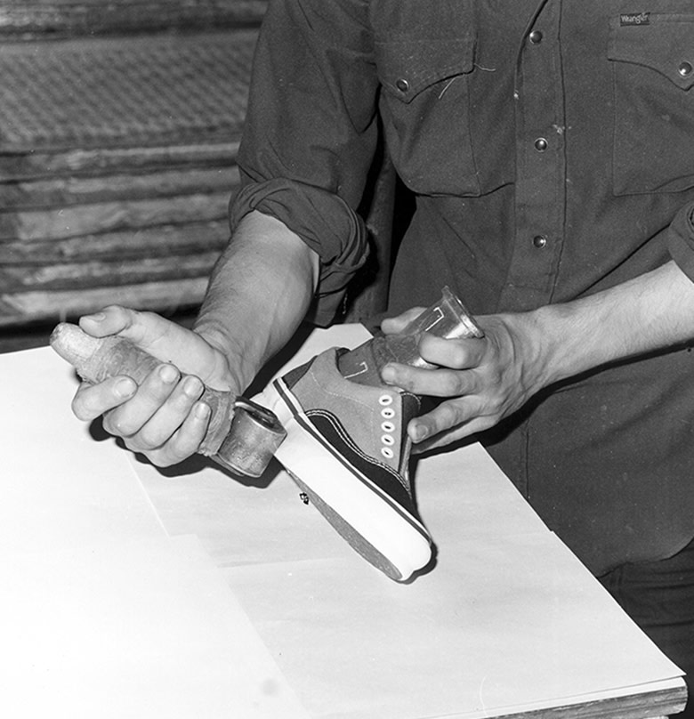 A black and white photo of a man making a custom Era shoe and using a roller to press on the rubber walls.