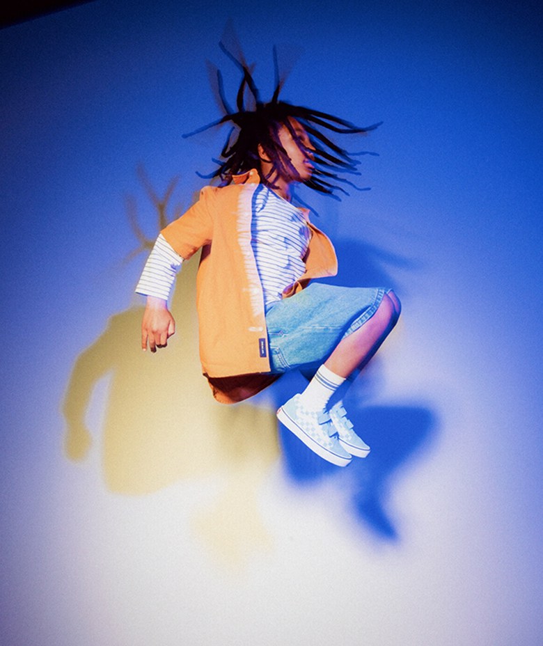 A kid jumping upwards in a blue room with a yellow shadow wearing a yellow overshirt, denim shorts, and Vans Customs shoes.