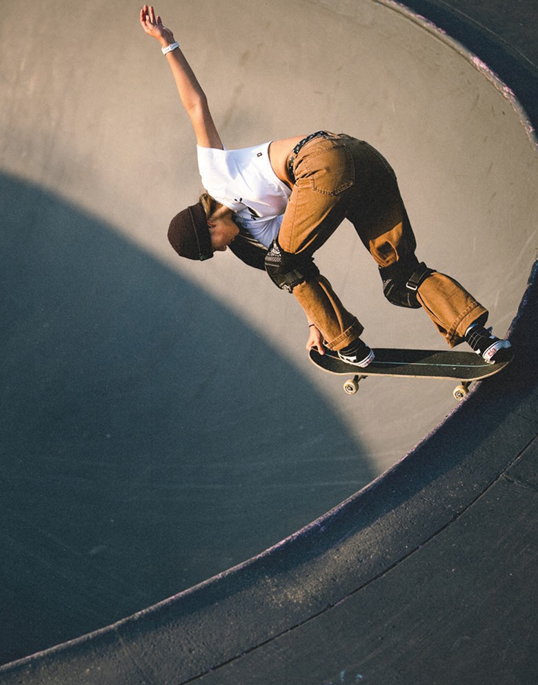 A skater grabs her board while on the lip of a bowl wearing a white t-shirt, brown pants, and black Sk8-Hi with a white SideStripe.