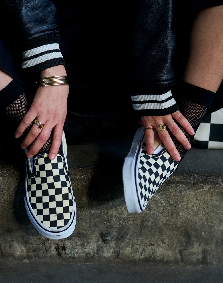 A person sits on a concrete steps with black and white Checkerboard Slip-On shoes.
