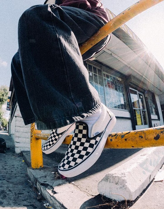 A person sitting on a yellow handrail wearing black and white Checkerboard Classic Slip-On outside of a convenience store.