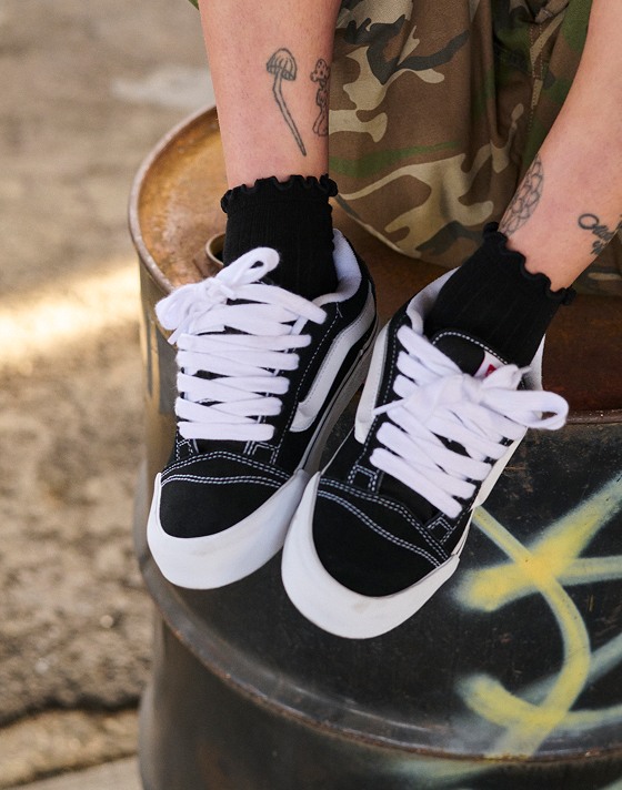 A person sitting on a metal barrel wearing black and white Knu Skool shoes with camo shorts.