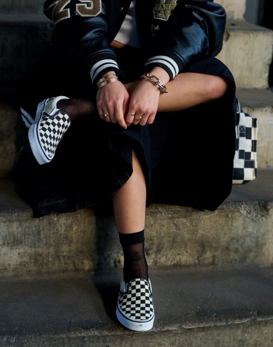 A person sits on a concrete steps with black and white Checkerboard Slip-On shoes.