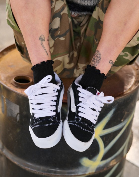 A person sitting on a metal barrel wearing black and white Knu Skool shoes with camo shorts.