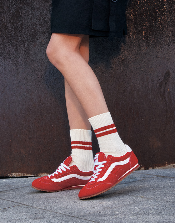 A person wearing red Vans Super Lowpro sneakers with white details and white socks with red stripes stands on a sidewalk. The person is dressed in a black outfit, and the background features a textured, dark brown wall.