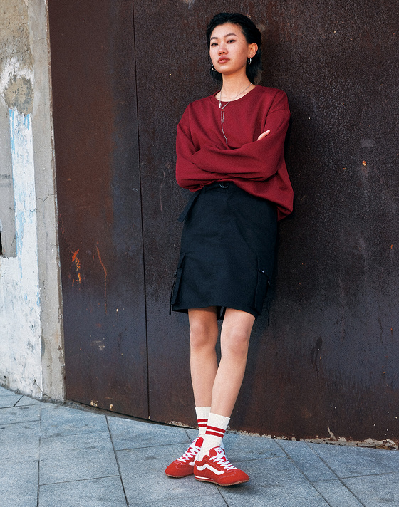 A person stands against a dark wall with arms crossed, wearing a red sweater, black skirt, red and white Vans Super Lowpro sneakers, and white socks. The background includes a textured brown and concrete wall.