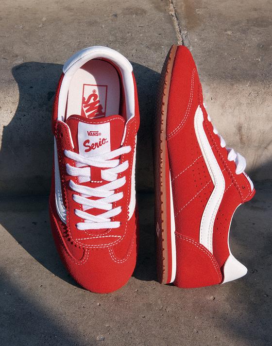 A pair of red Vans Super Lowpro shoes is displayed on concrete steps. One shoe is upright, showing white laces and a white stripe logo. The other is on its side, highlighting the gum sole and Vans branding on the insole and tongue.