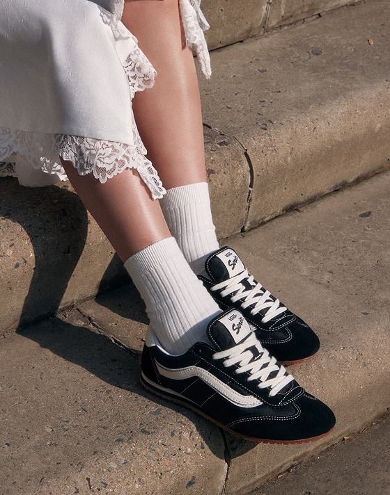 Person wearing a white lace-trimmed dress sits on steps, showcasing black and white Vans Super Lowpro sneakers paired with white ribbed socks. The scene is sunlit, highlighting the texture of the dress and shoes.