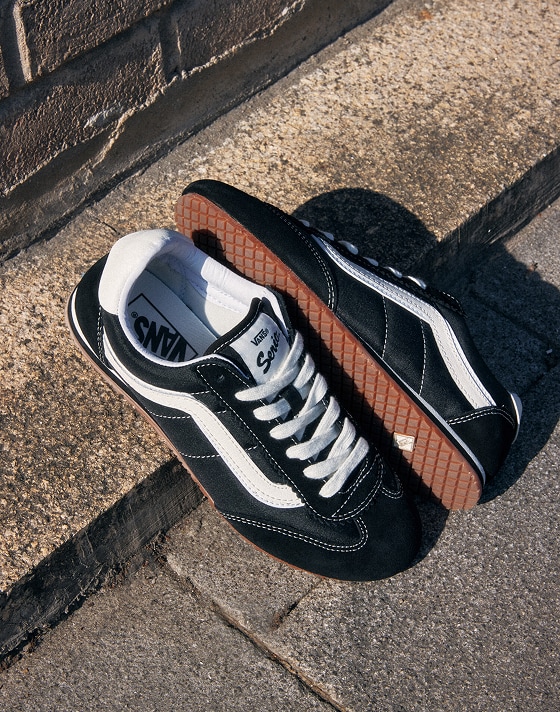 Black and white Vans Super Lowpro sneakers with Vans logo on a concrete surface. The shoes have suede details, white laces, and gum soles. They are positioned with one shoe upright and the other resting on its side, near a brick wall.