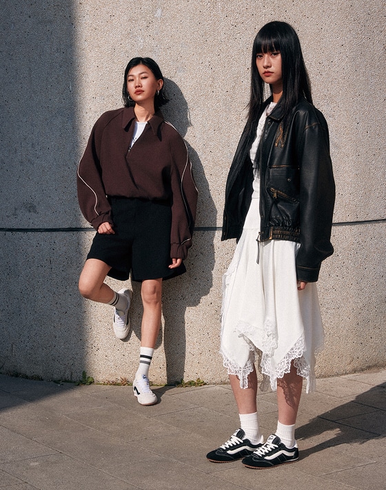 Two women stand outdoors against a concrete wall. One wears a brown jacket, black shorts, and white sneakers; she leans against the wall. The other wears a black leather jacket over a white dress with black Vans Super Lowpro sneakers and stands facing forward.