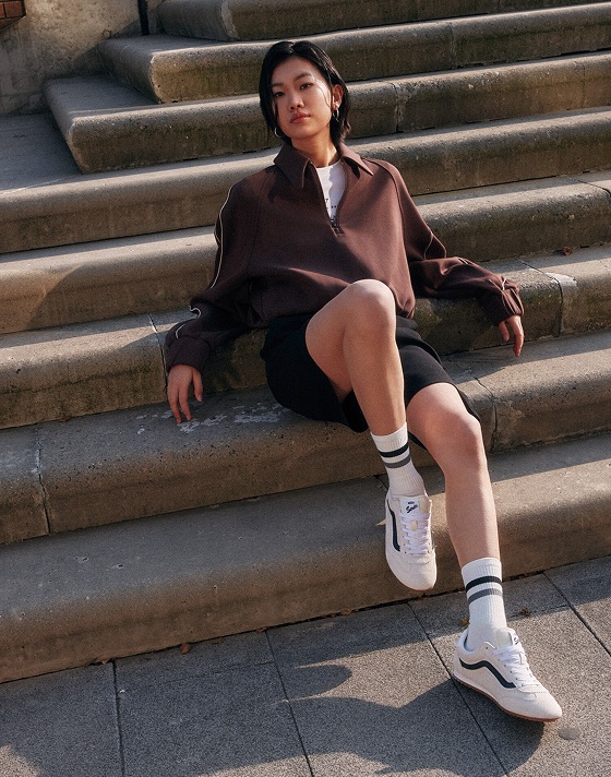 A person sits casually on stone steps wearing a brown long-sleeve shirt, black shorts, striped socks, and white Vans Super Lowpro shoes. They lean back, supporting themselves with one arm, gazing into the distance outdoors.