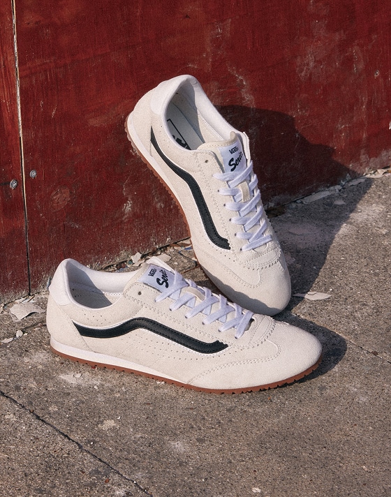 A pair of white Vans Super Lowpro sneakers with black stripes on the side and white laces is placed on a rough, textured concrete surface. The background features a weathered, red-painted wooden panel.