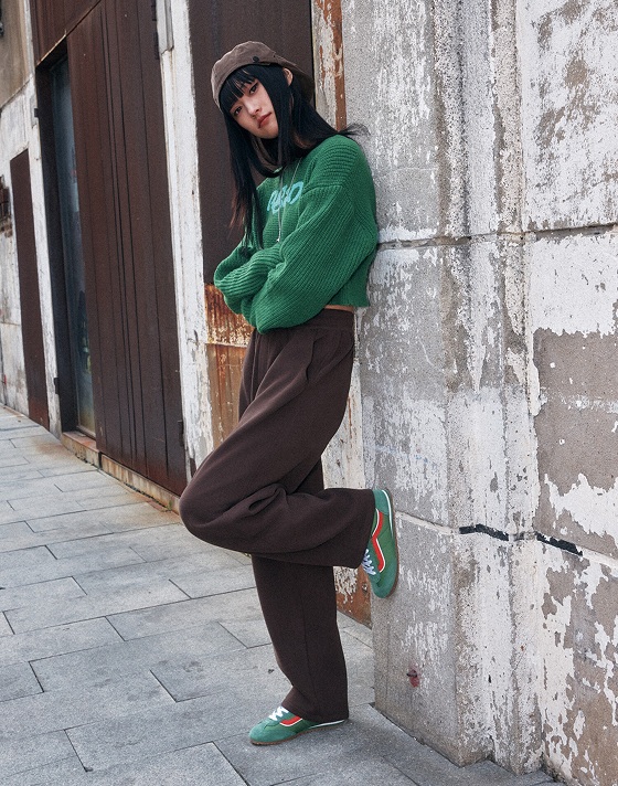 A person with long dark hair leans against a weathered wall in an urban setting. They wear a green sweater, brown pants, green Vans Super Lowpro sneakers, and a brown cap. One foot is propped against the wall and their arms are crossed.