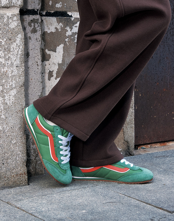 A person leans against a textured wall on a city street, wearing brown pants and green Vans Super Lowpro sneakers with white and red stripes. The focus is on the lower half, highlighting the shoes and casual stance.
