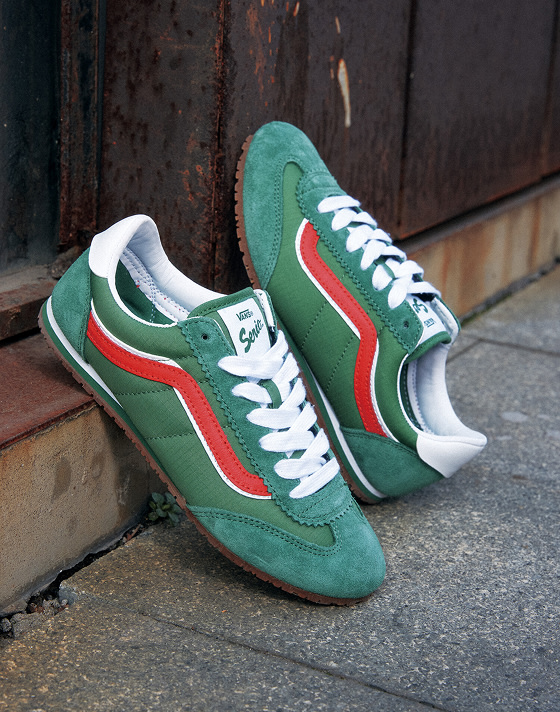 A pair of green Vans Super Lowpro sneakers with white laces, white accents, and red stripes sits on a concrete surface against a rusted metal background.