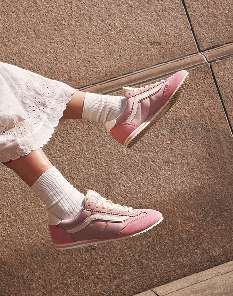 A person wearing Sepia Rose Vans Super Lowpro sneakers with white socks against a stone wall backdrop. They are wearing a white, lacy skirt. The image captures a dynamic mid-air pose. 