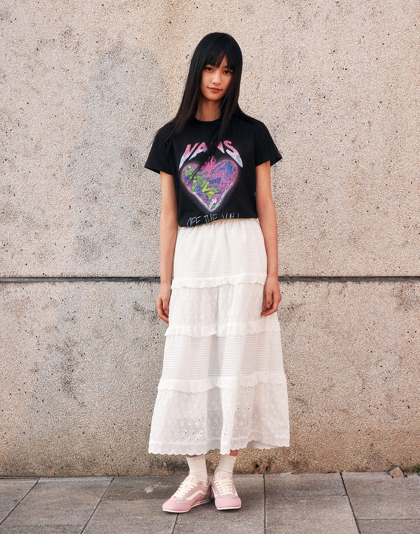 A person with long dark hair stands against a neutral textured wall. They wear a black Vans graphic T-shirt, a white tiered skirt, and Sepia Rose Vans Super Lowpro sneakers with white socks. Their arms are relaxed by their sides, and they look directly at the camera.
